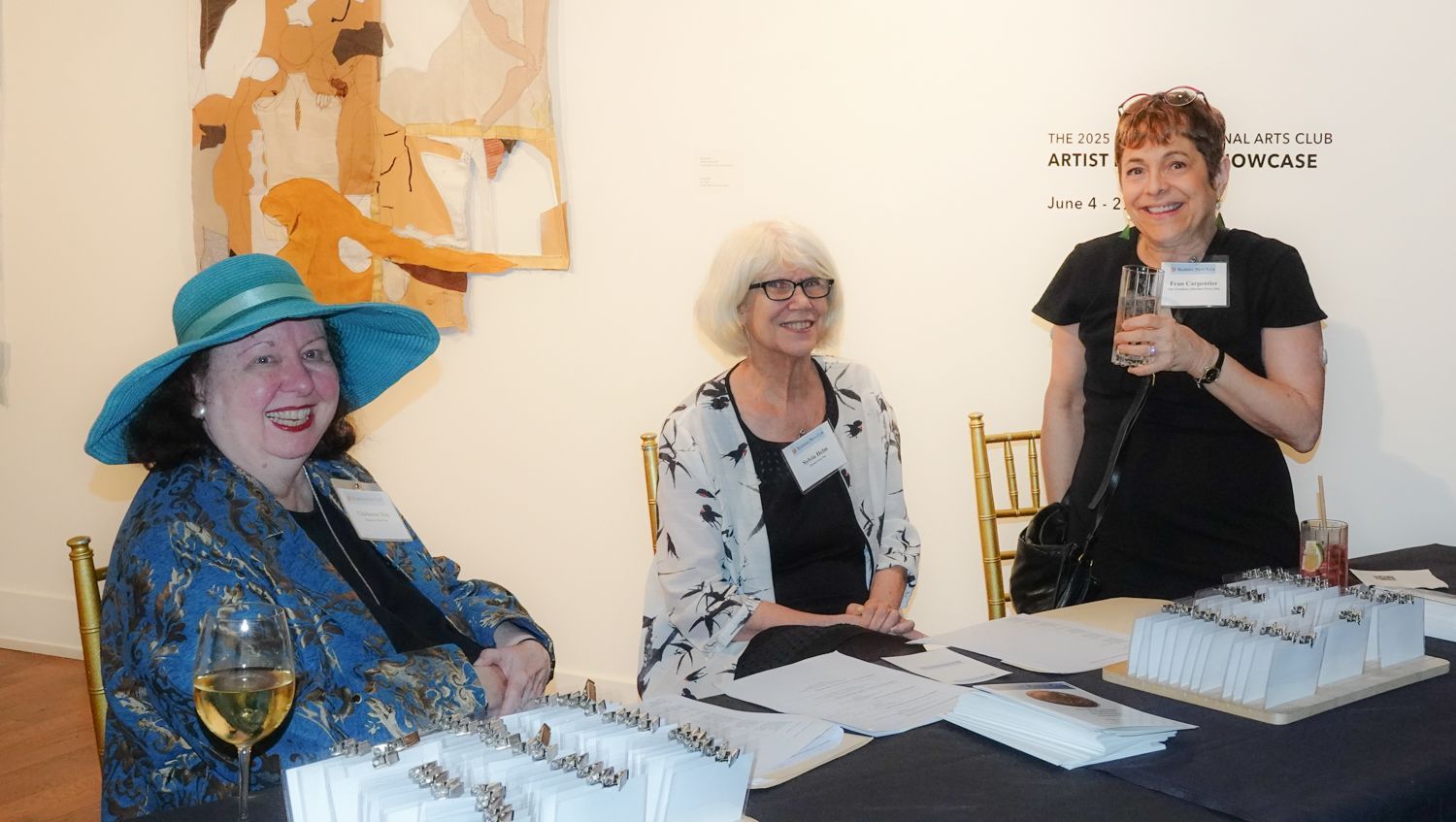 Three women at a table with name tags, smiling. One wears a blue hat and jacket, another a floral shirt, the third holds a microphone.