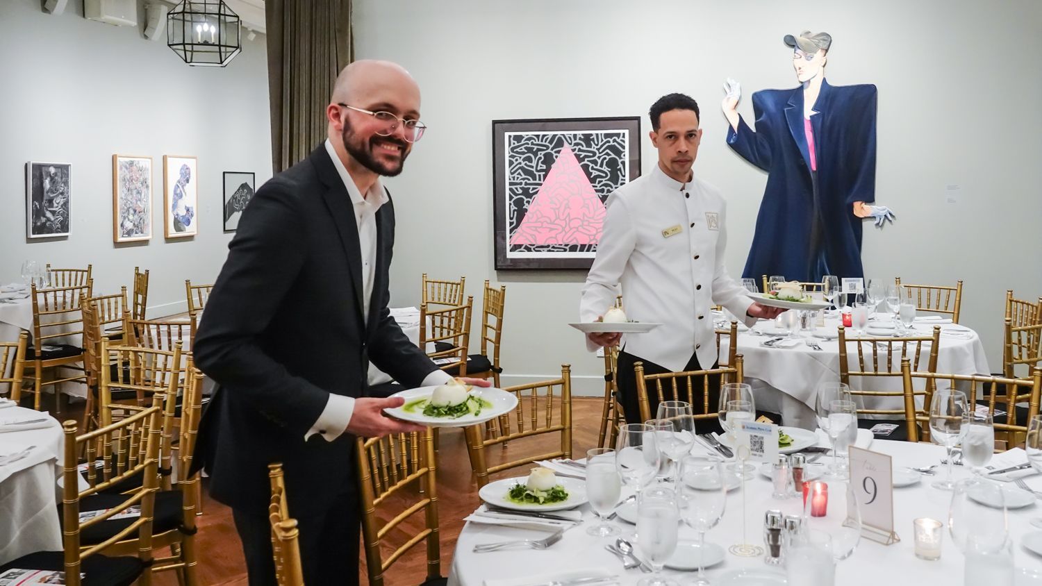 Two servers setting tables in a dining room with artwork. One smiles, carrying a plate. Tables are set for a formal meal.