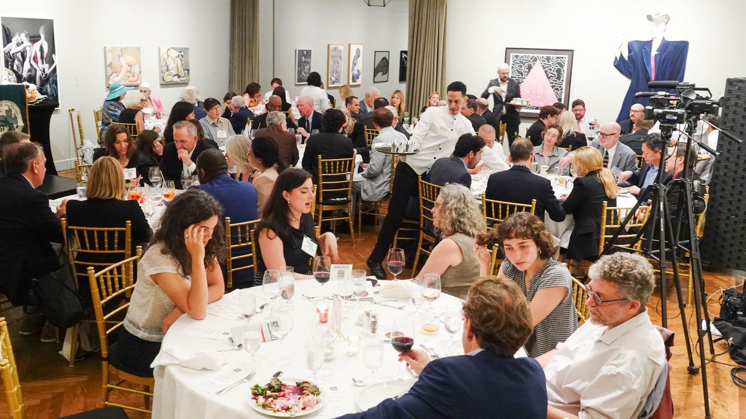 People at round tables in a gallery, attending an event with food, wine, and artwork on the walls.