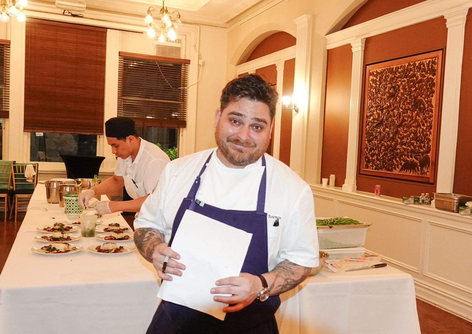 Chef in a white shirt and blue apron smiling, holding paper at a table with food.