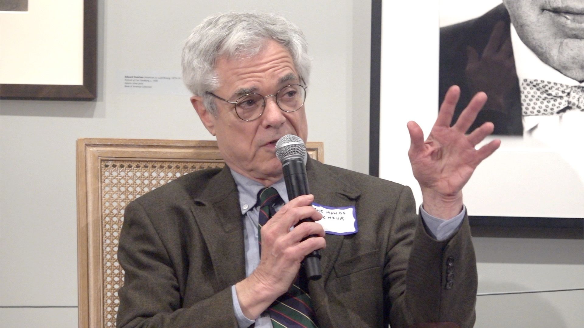 Man in glasses speaks into microphone, gesturing with right hand. He wears a jacket and shirt, seated in a room with art.