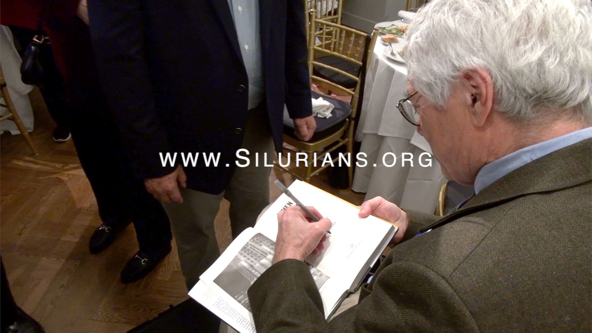Man signing a book. Others nearby. Indoors, book open.