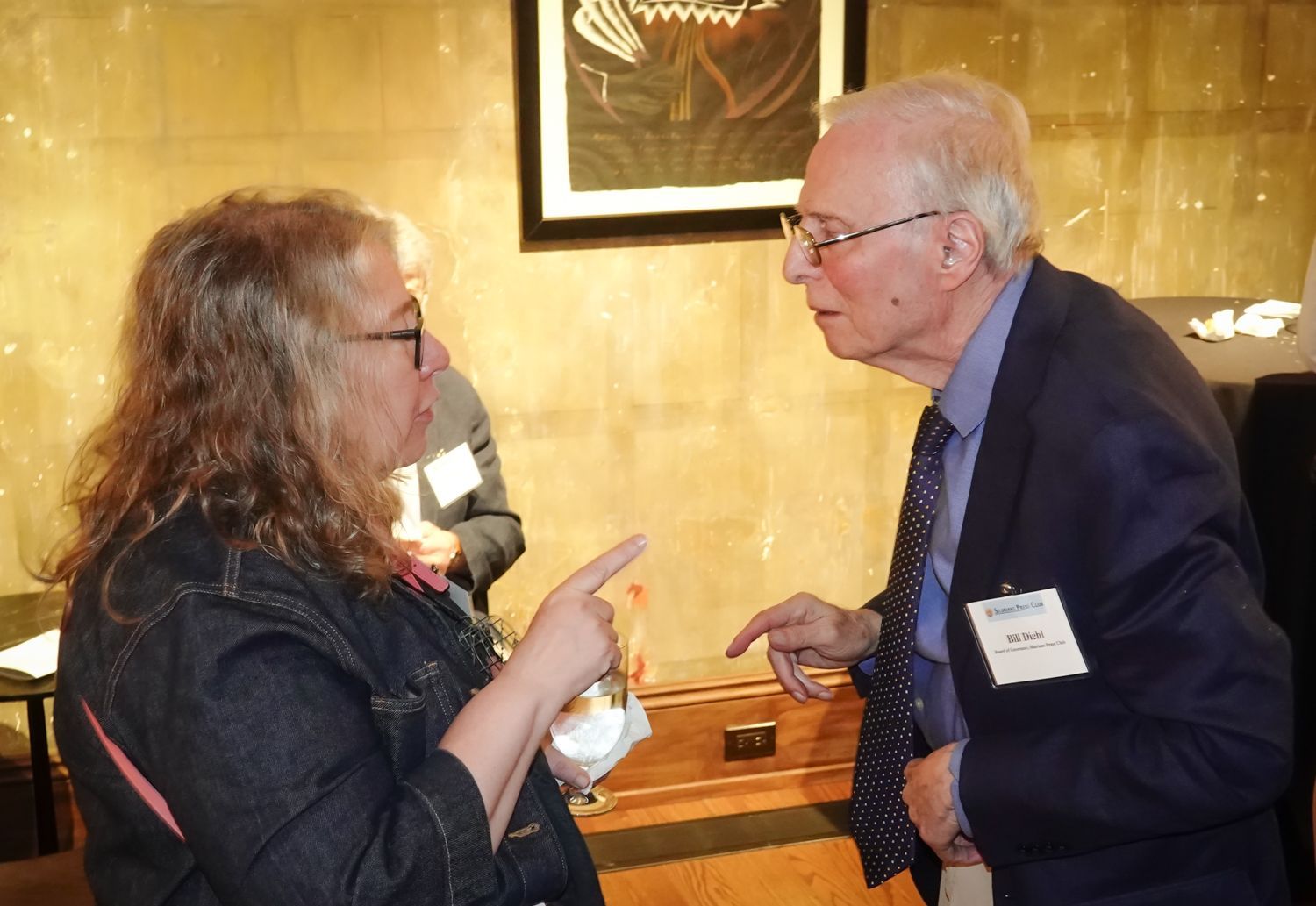 Woman and man in conversation, pointing. Woman wears denim jacket; man wears blazer. Indoor setting, artwork in background.