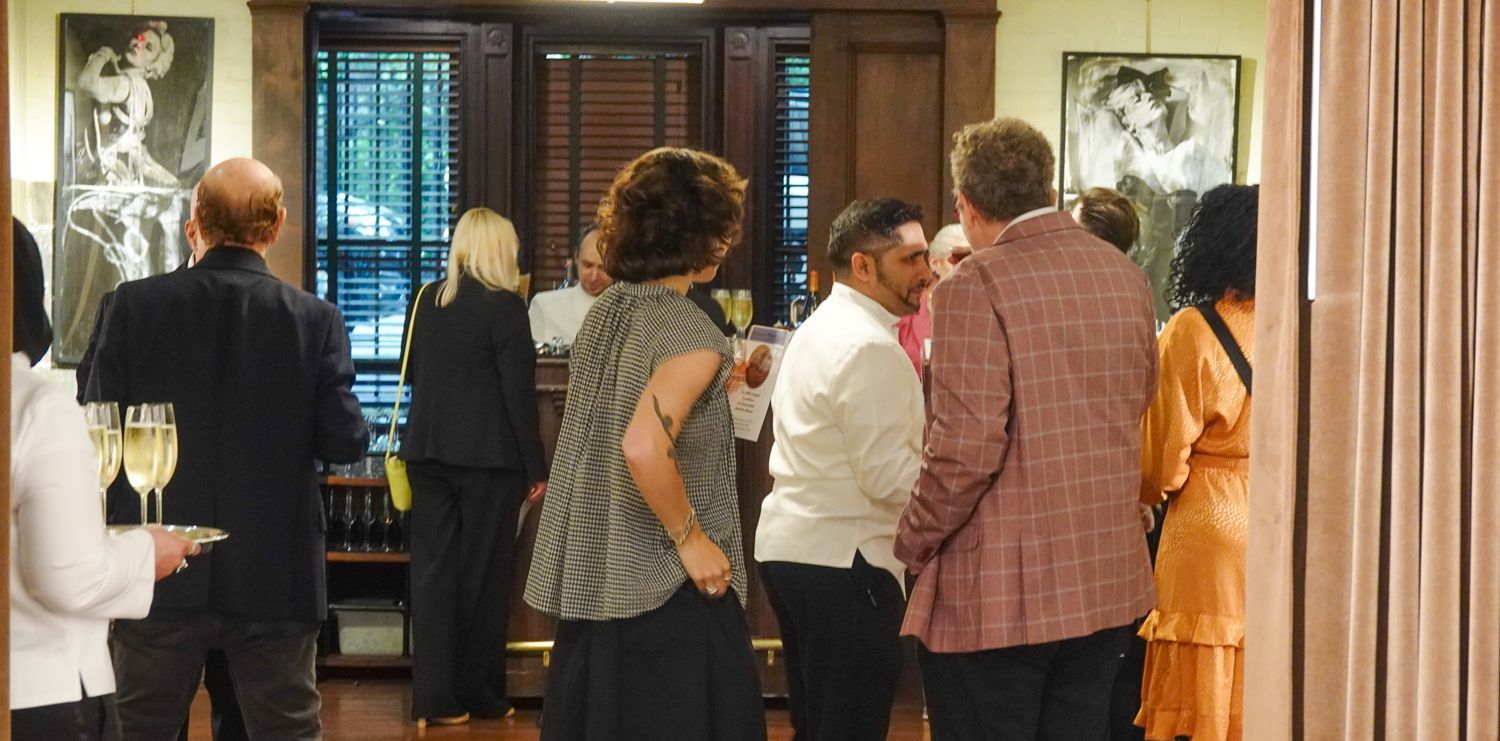 People in a room, some holding drinks. There is art on the walls, and the room has a wooden decor.