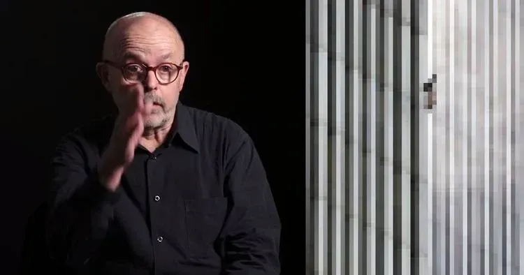 A bald man in glasses gestures with one hand against a dark background beside vertical white blinds.
