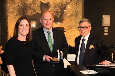 Three people at a formal event; man in suit holds medal, two others beside him, gold artwork background.