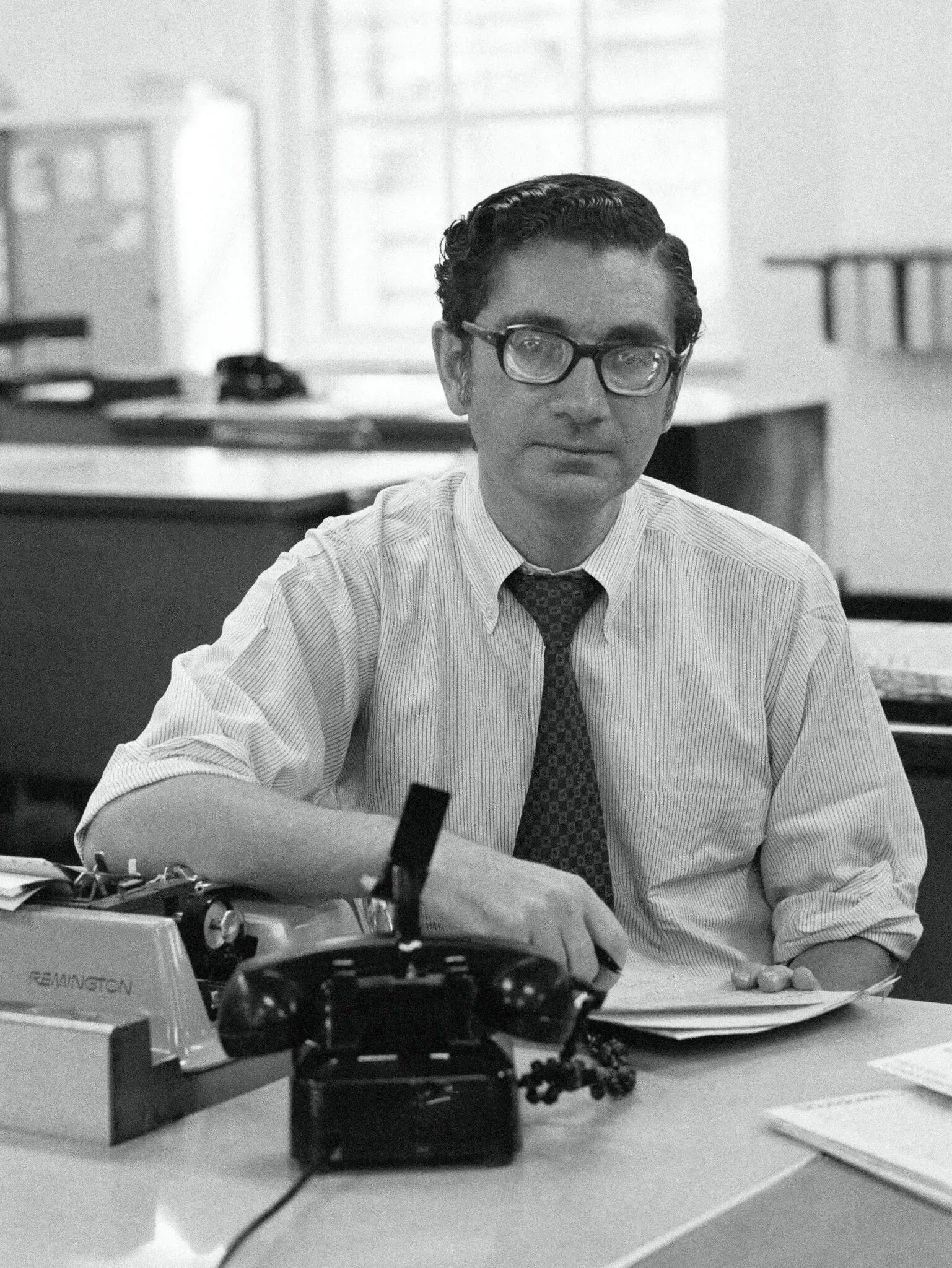 Man wearing glasses and tie sits at desk with phone and typewriter in a bright office.