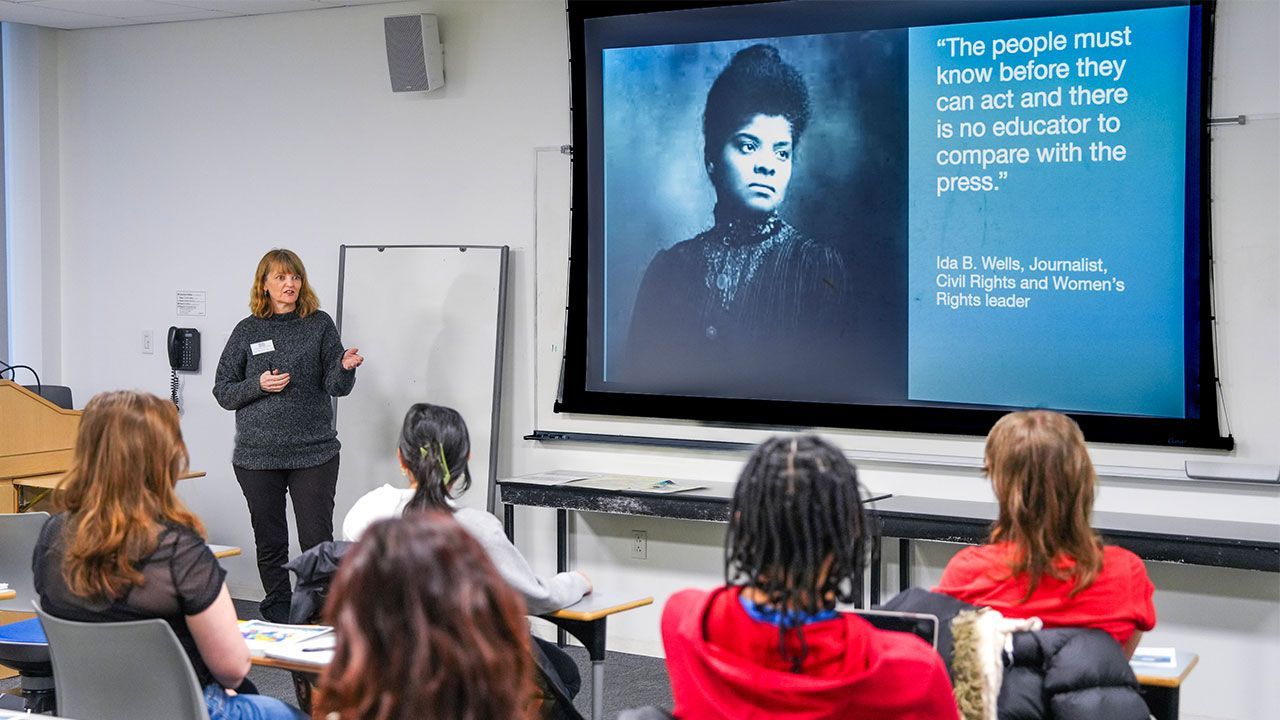 A presenter in a classroom gives a lecture with a projector screen showing a woman and a quote.