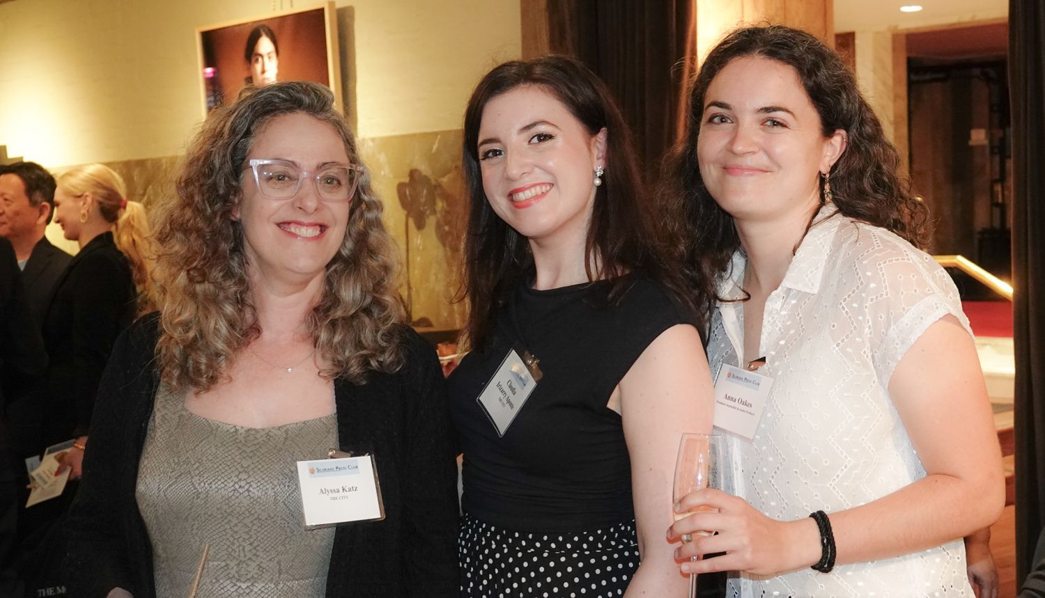 Three women smiling at an event. The woman on the left has glasses and graying curly hair.