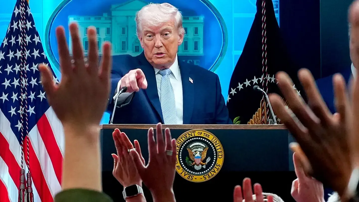 Donald Trump speaking at a podium with U.S. flags behind him as audience hands are raised in front