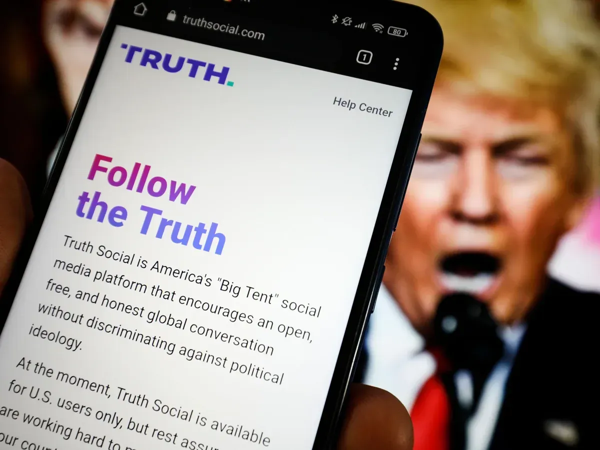 Phone displaying “Follow the Truth” beside blurred Trump speech in background