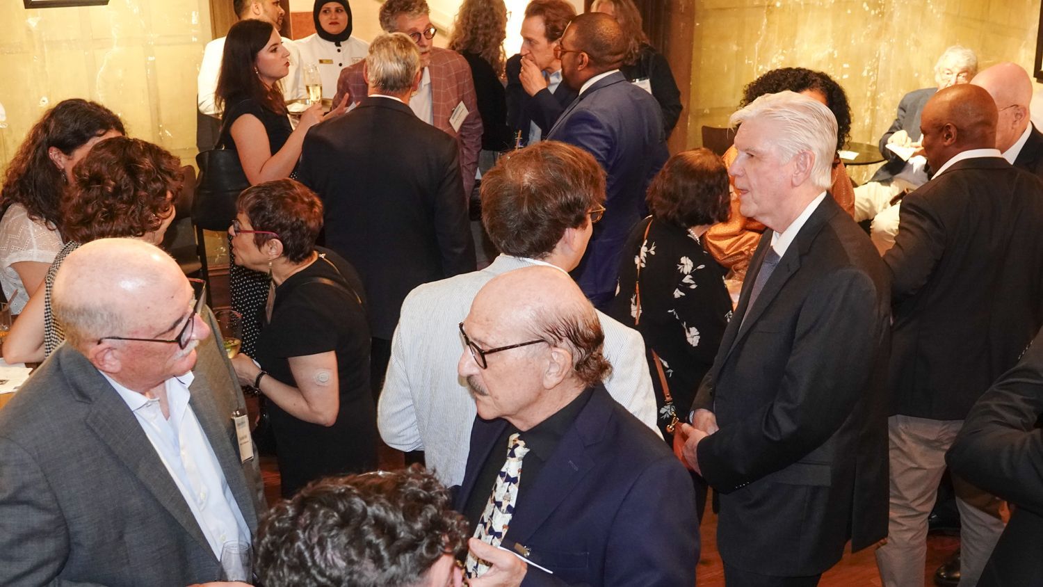 People in suits and other attire mingle and converse in a crowded interior setting.