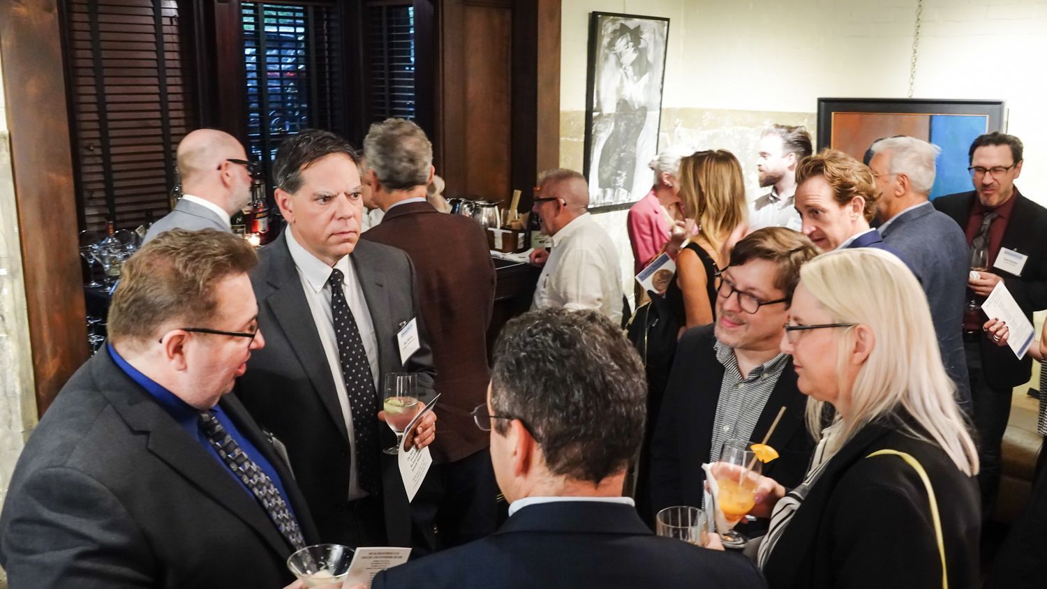 People in suits and dresses mingle at an event, some holding drinks. Artwork and dark wood in the background.