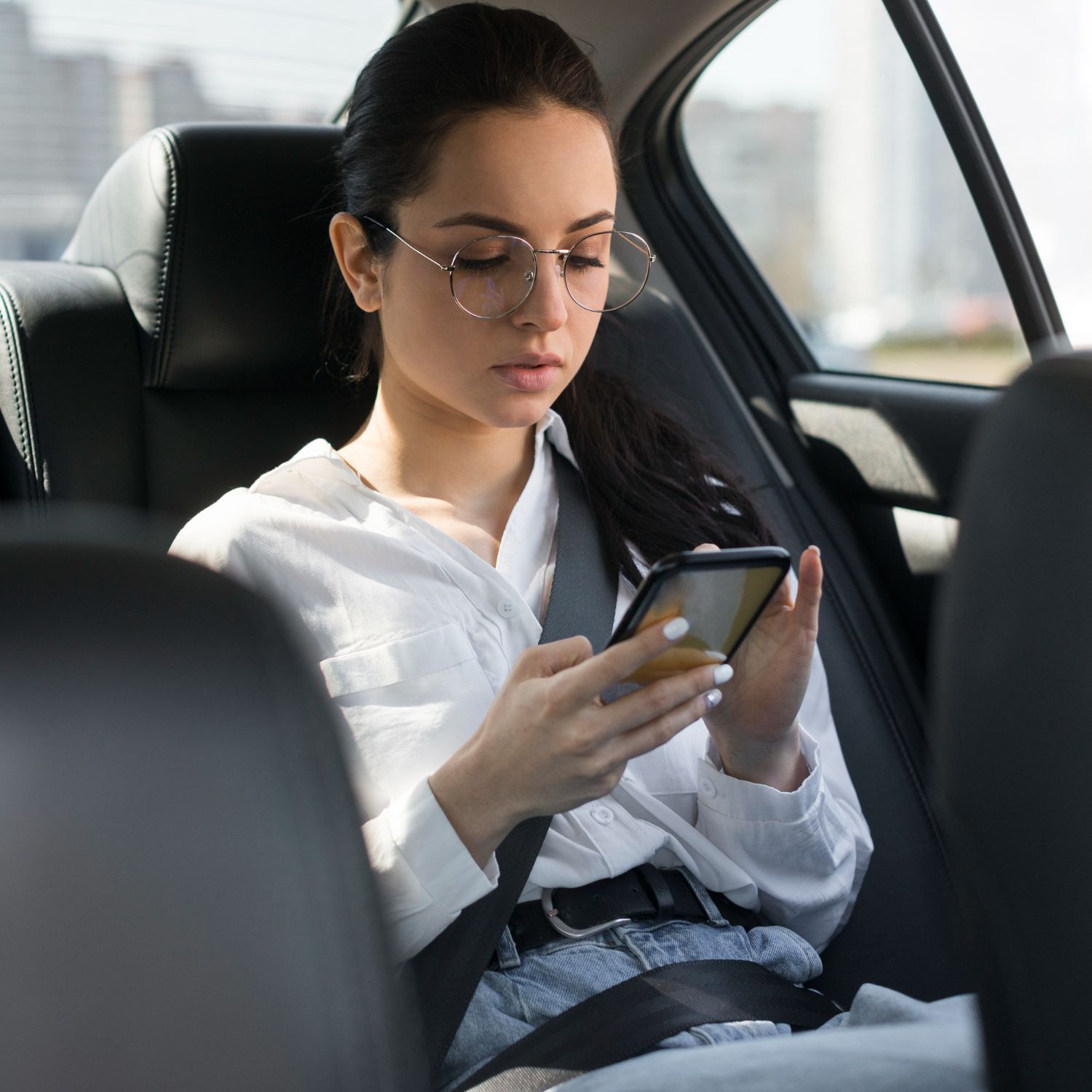Woman in car, wearing glasses, looking at phone, seatbelt on. 