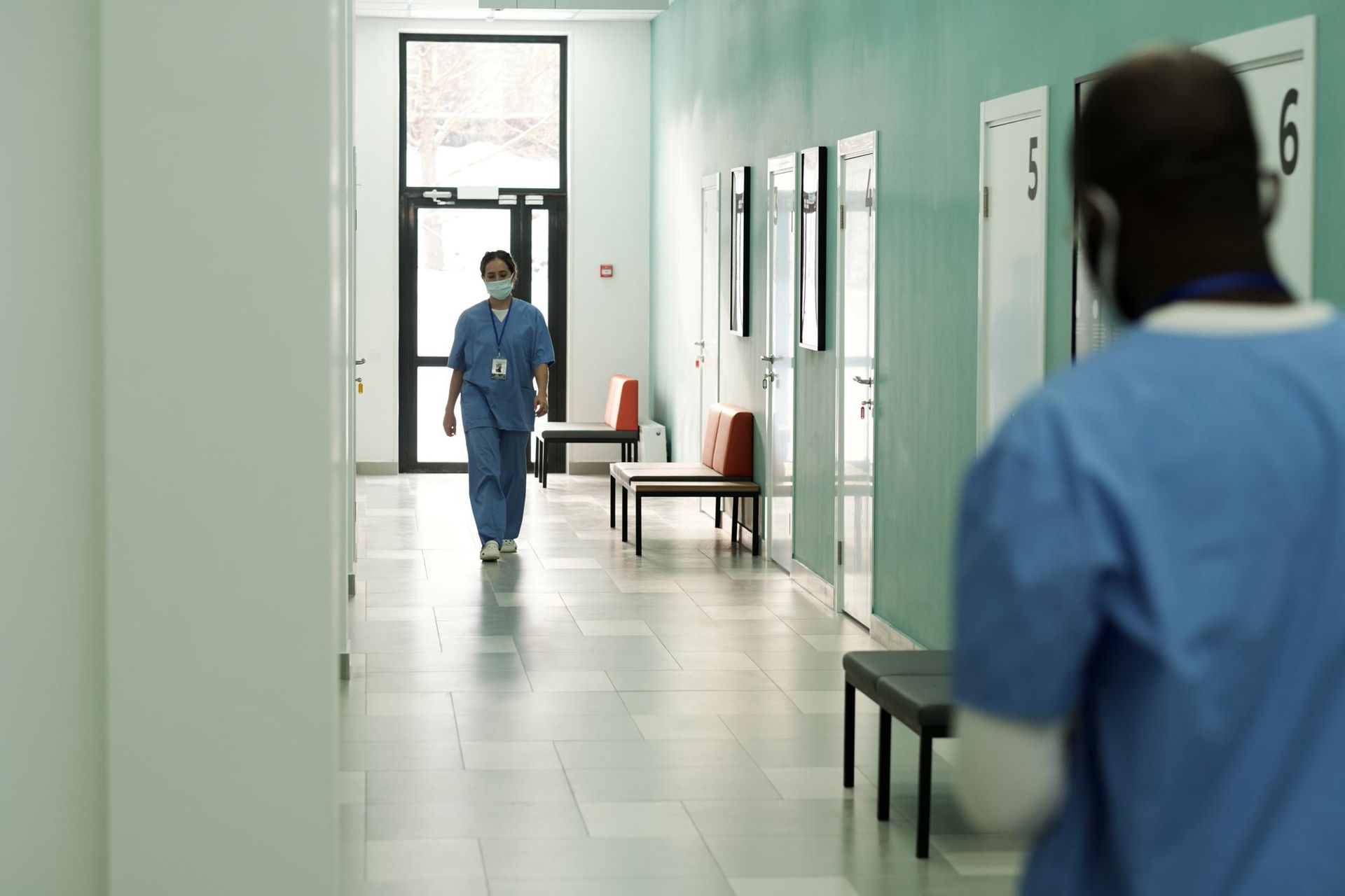 Nurse walking down a hospital hallway, another person in scrubs is in the foreground.