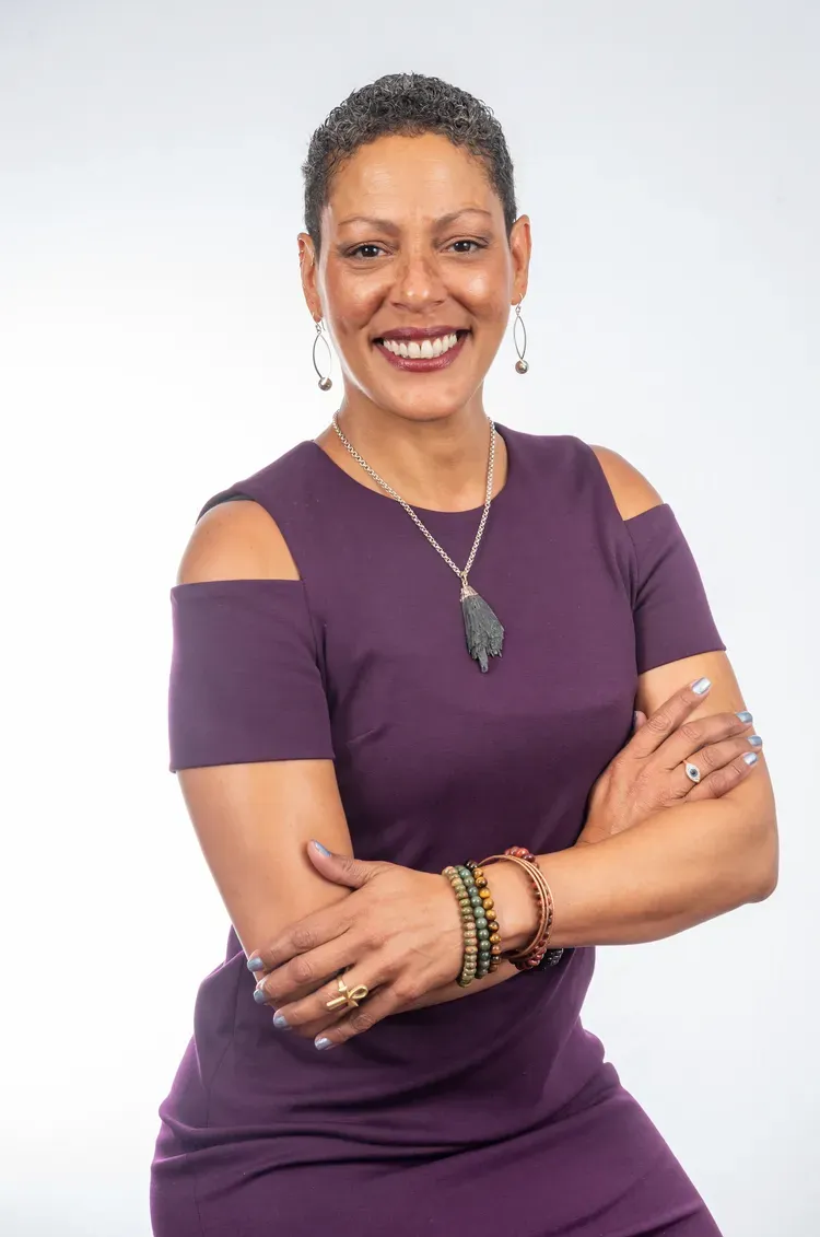 Woman with short hair, smiling, arms crossed, wearing a purple dress and jewelry, standing in front of a white background.