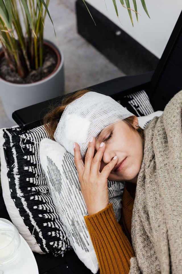 Person resting on a couch with a bandaged head and hand over face, wearing a blanket, near a potted plant.