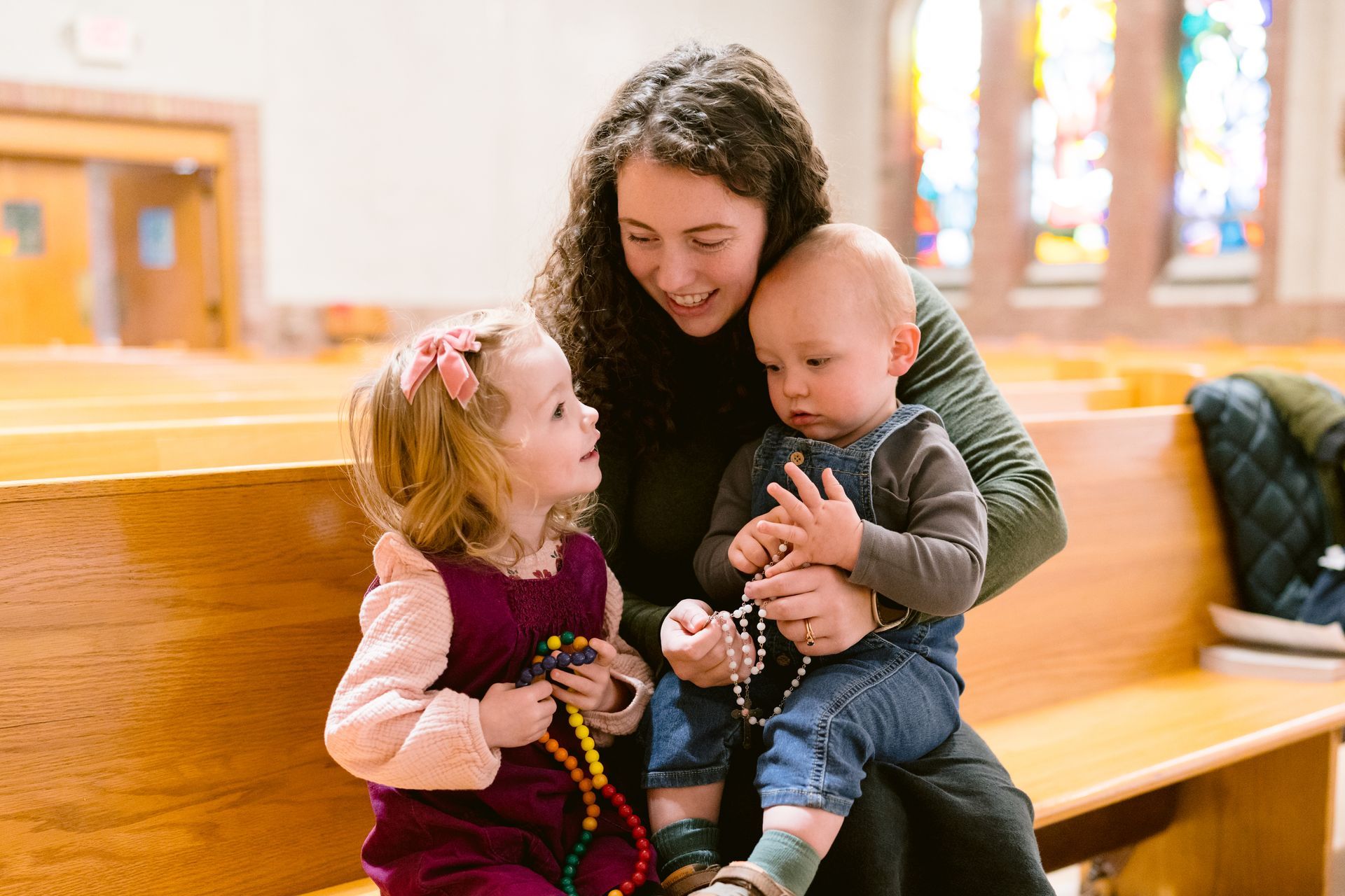 Woman sitting with two children in a church pew, sharing rosary beads and smiling.
