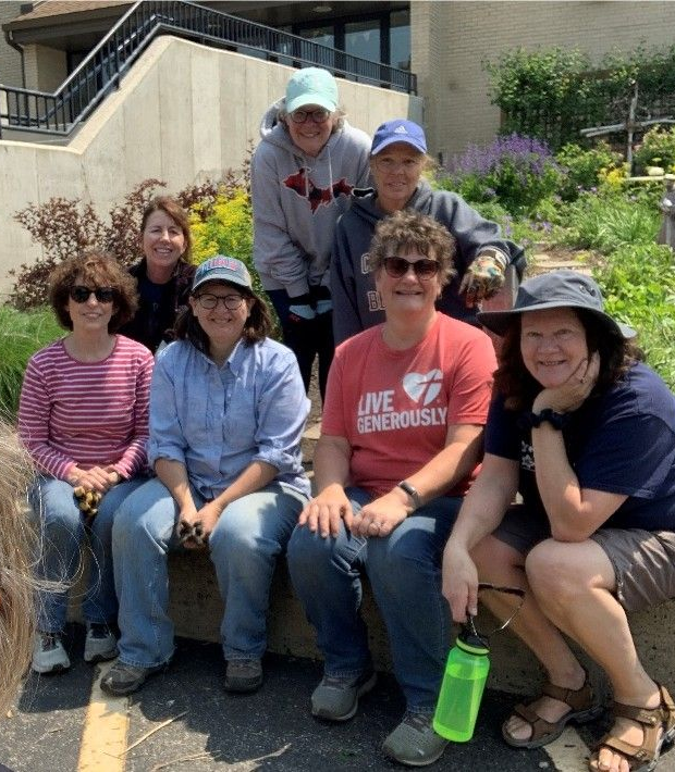 Six people sitting outdoors on a bench in a garden, smiling at the camera.