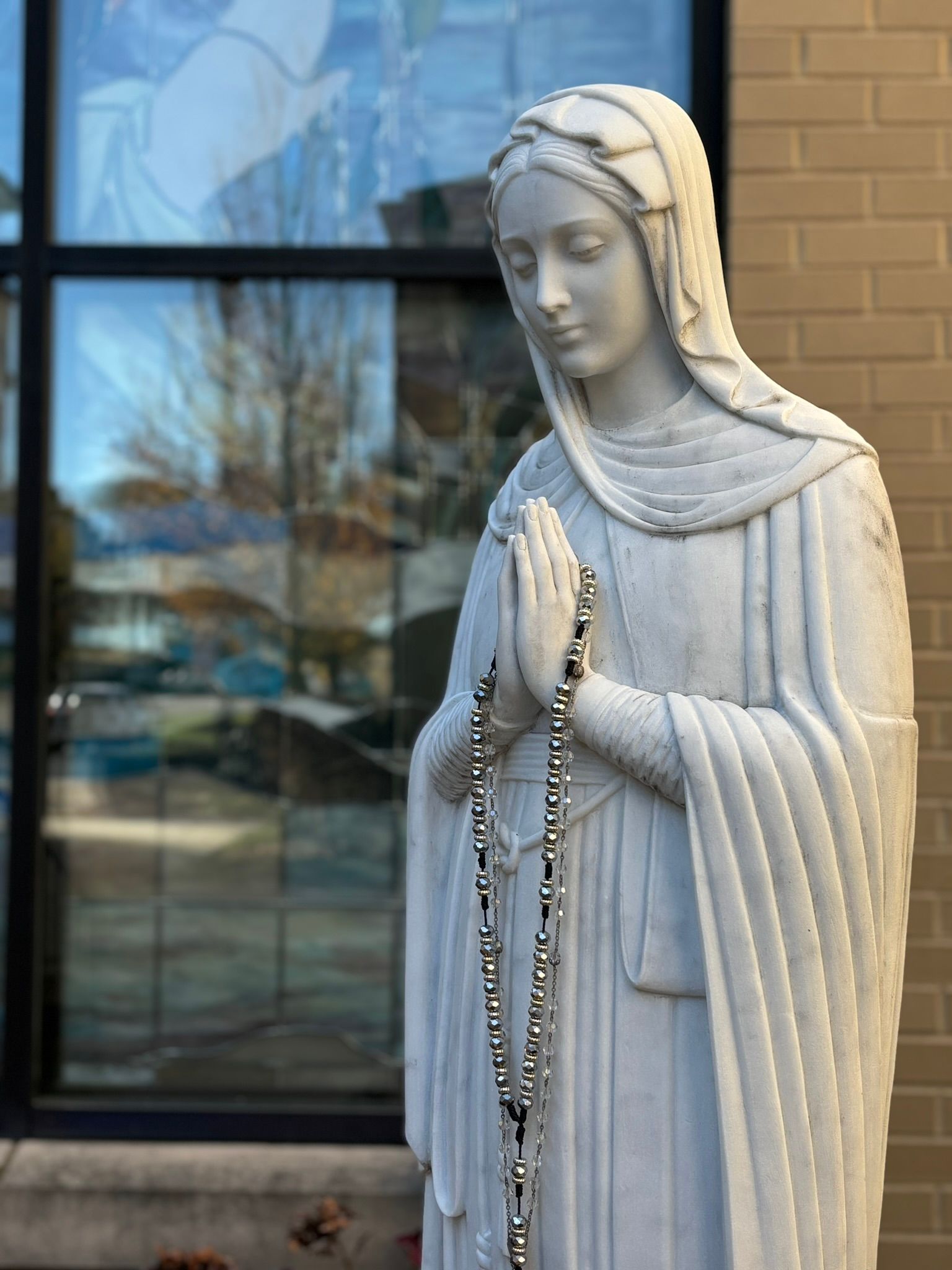 White statue of a robed figure holding rosary beads outside a building window