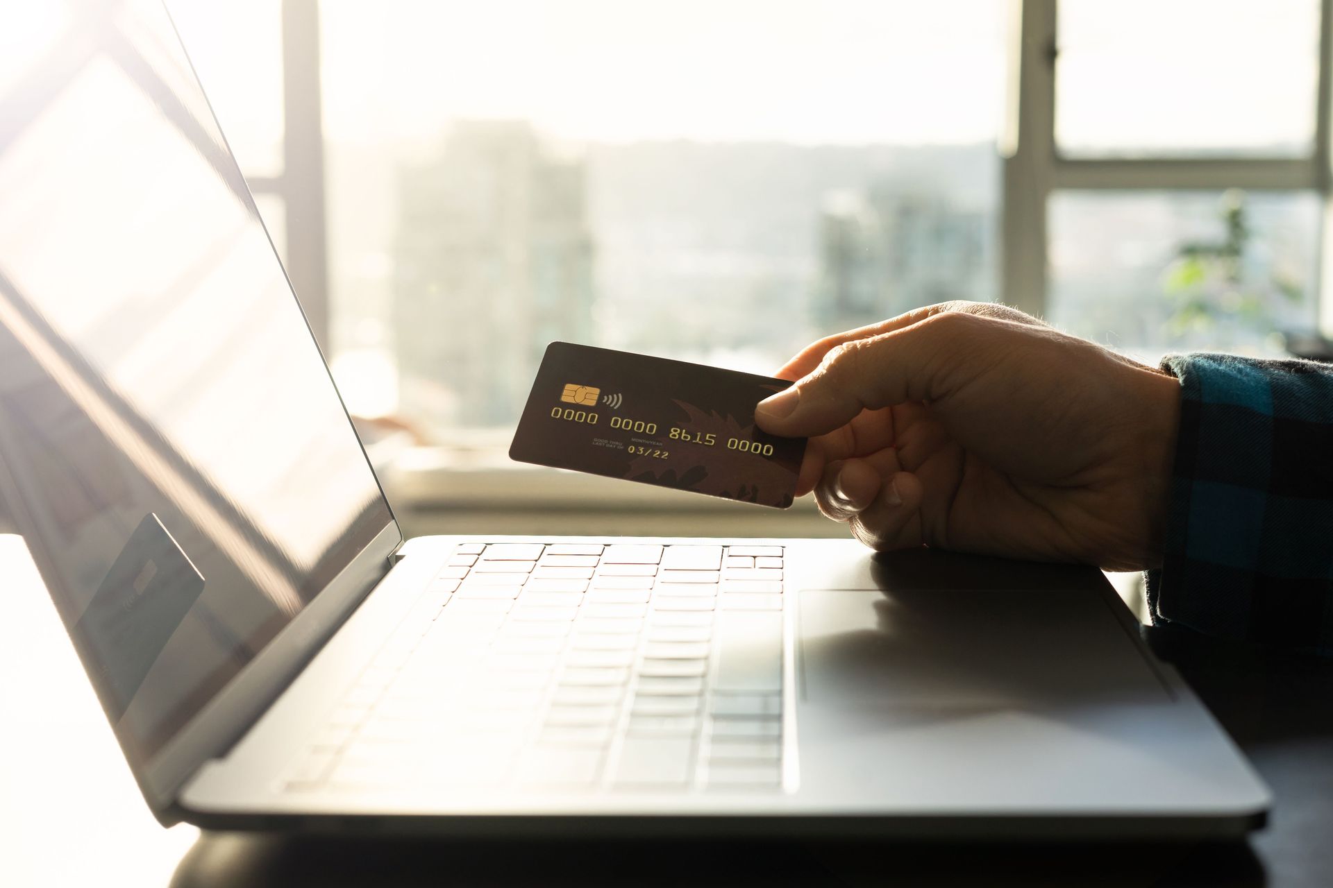 Hand holding a credit card near a laptop for online shopping on a bright desk