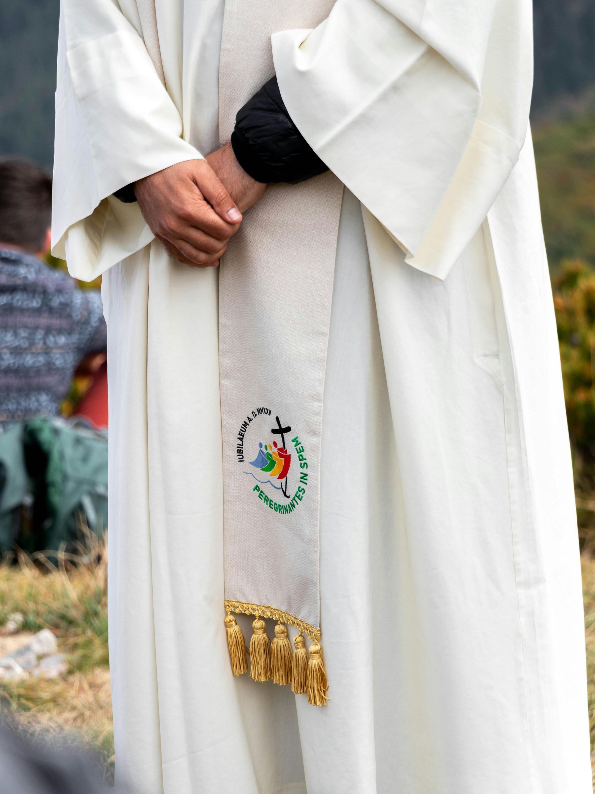 Person in a white robe standing outdoors, hands clasped, with an embroidered stole hanging down.