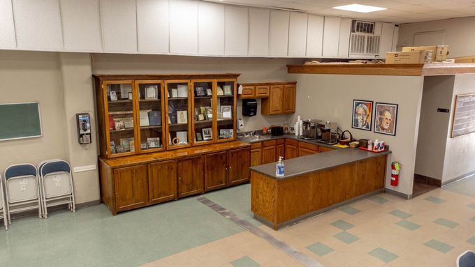 A room with a large wooden cabinet, kitchen area, and folding chairs. Beige and green colors are present.