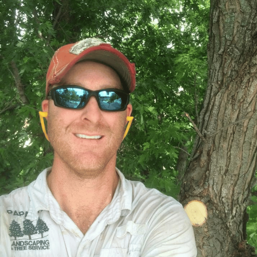 Smiling person in sunglasses and a red cap standing beside a tree in a green wooded area