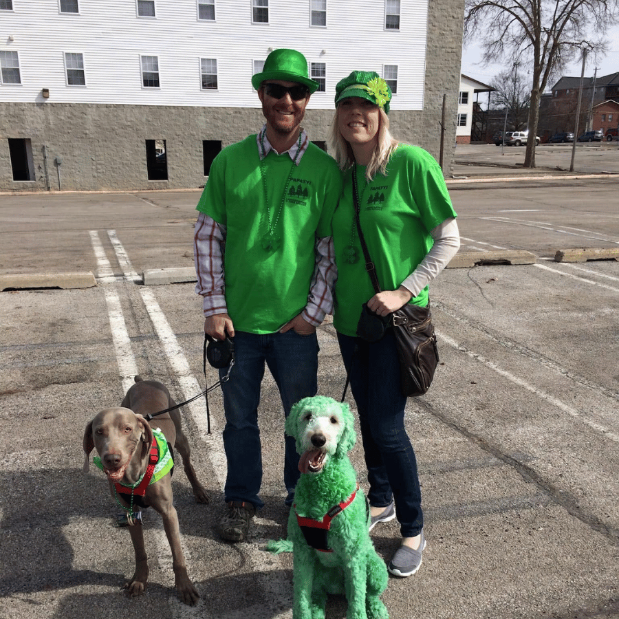 Two people in green shirts stand with two leashed dogs in a parking lot.