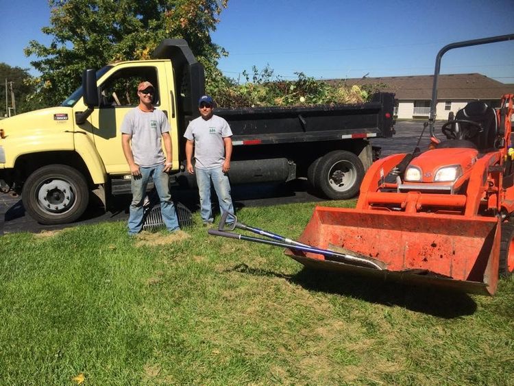 Two men stand beside a yellow truck and orange tractor on a grassy field.