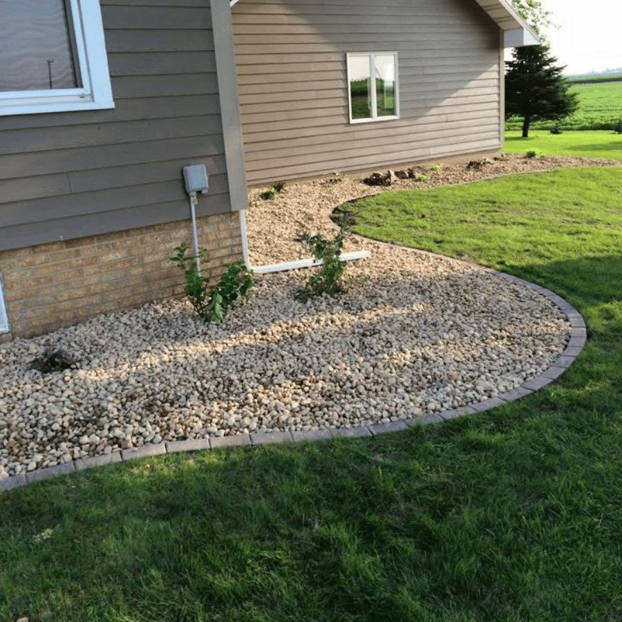 House with brown siding, bordered by a rock garden and green lawn, under a clear sky. A small tree stands nearby.