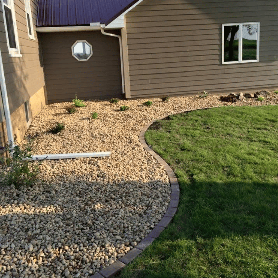 A house with a brown exterior features a landscaped yard with gravel and a curved brick border separating grass and stones.