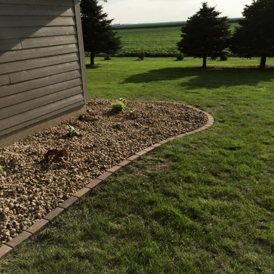 A garden bed with rocks and young plants borders a wooden house, surrounded by a lush green lawn and distant trees.