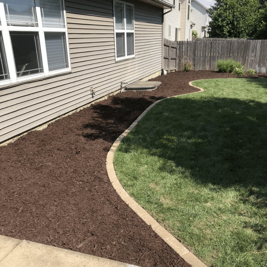 Curved garden bed with fresh mulch lines a house, bordered by green grass and a wooden fence under a clear blue sky.