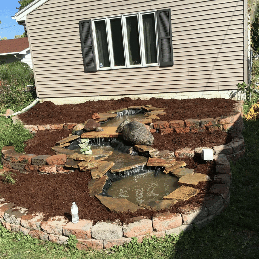 A small backyard waterfall flows over rocks into a pond, surrounded by mulch and a brick border near a house with beige siding.