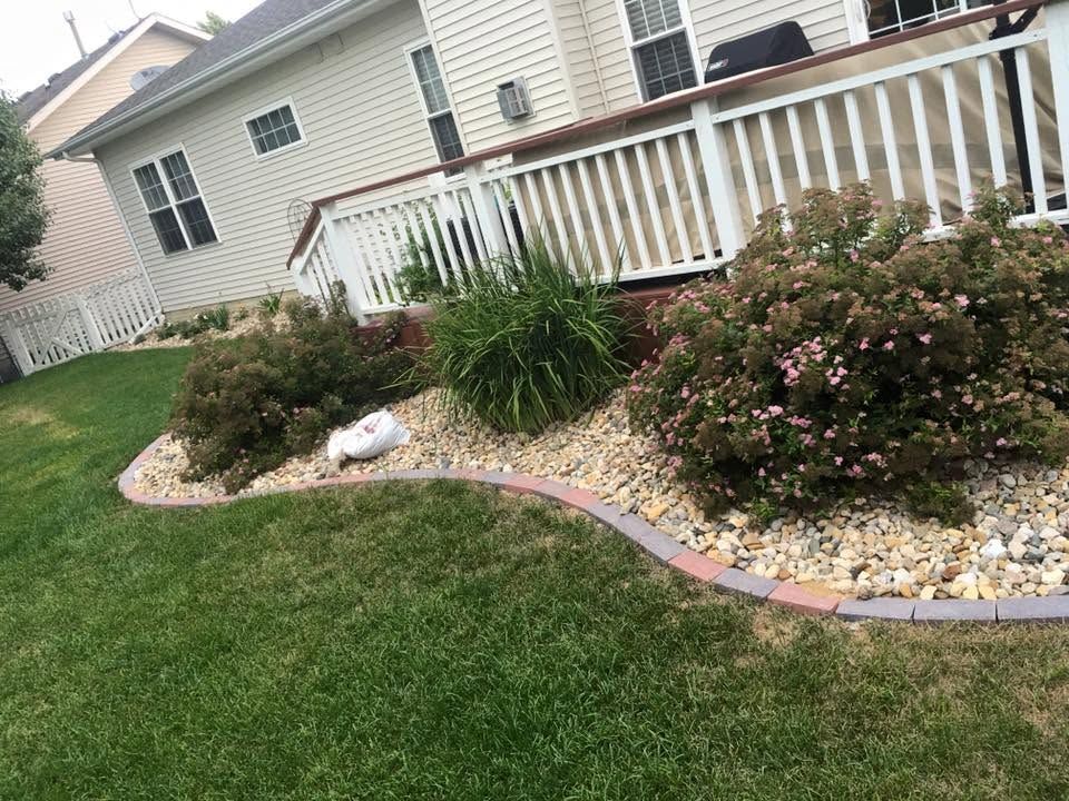 Curved flower bed with shrubs and pink blossoms beside a grassy yard and white porch railing