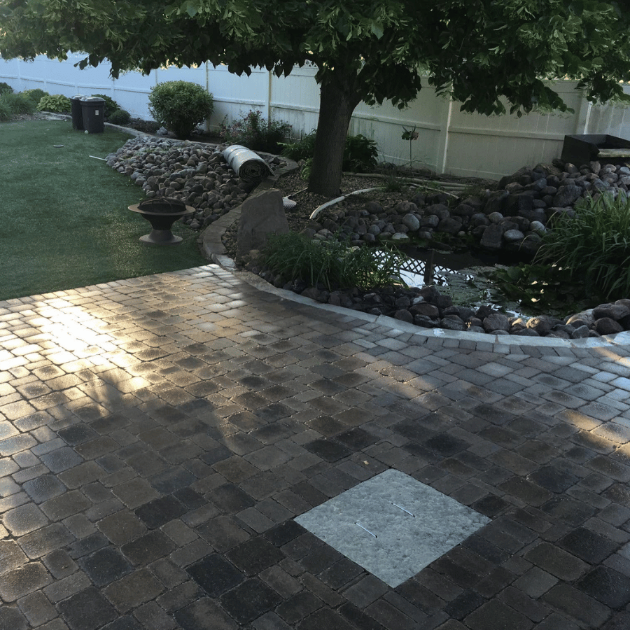Backyard patio with brick pavers, tree shade, and a landscaped rock border around a garden bed.