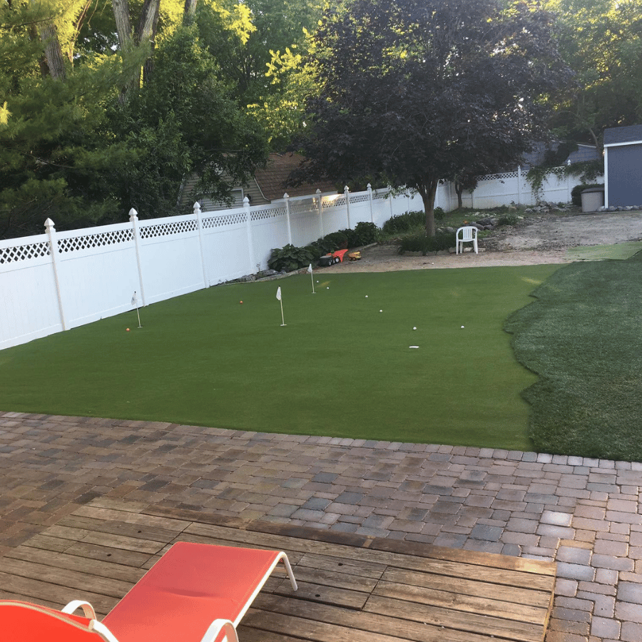 Backyard with white fence, green artificial turf, and a wooden deck with a red lounge chair.