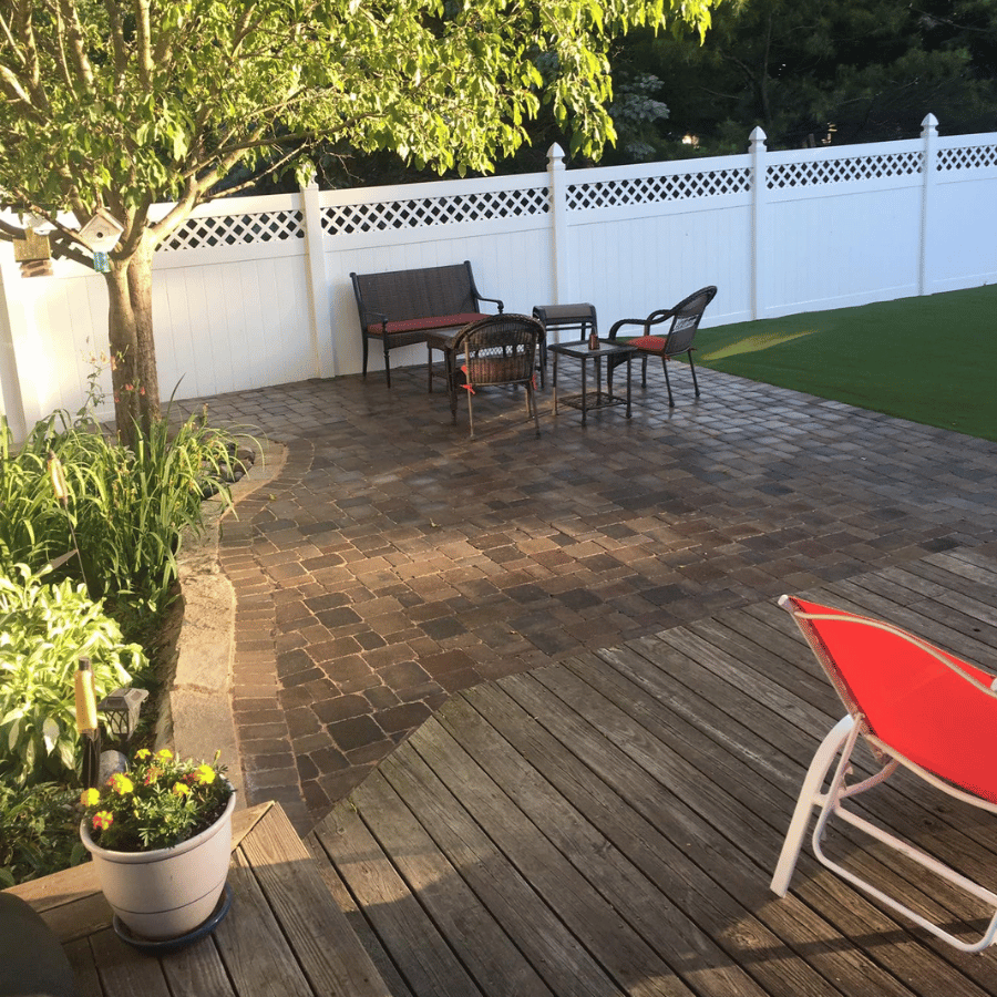 Backyard patio with wood deck, brick seating area, red lounge chair, table, and white fence