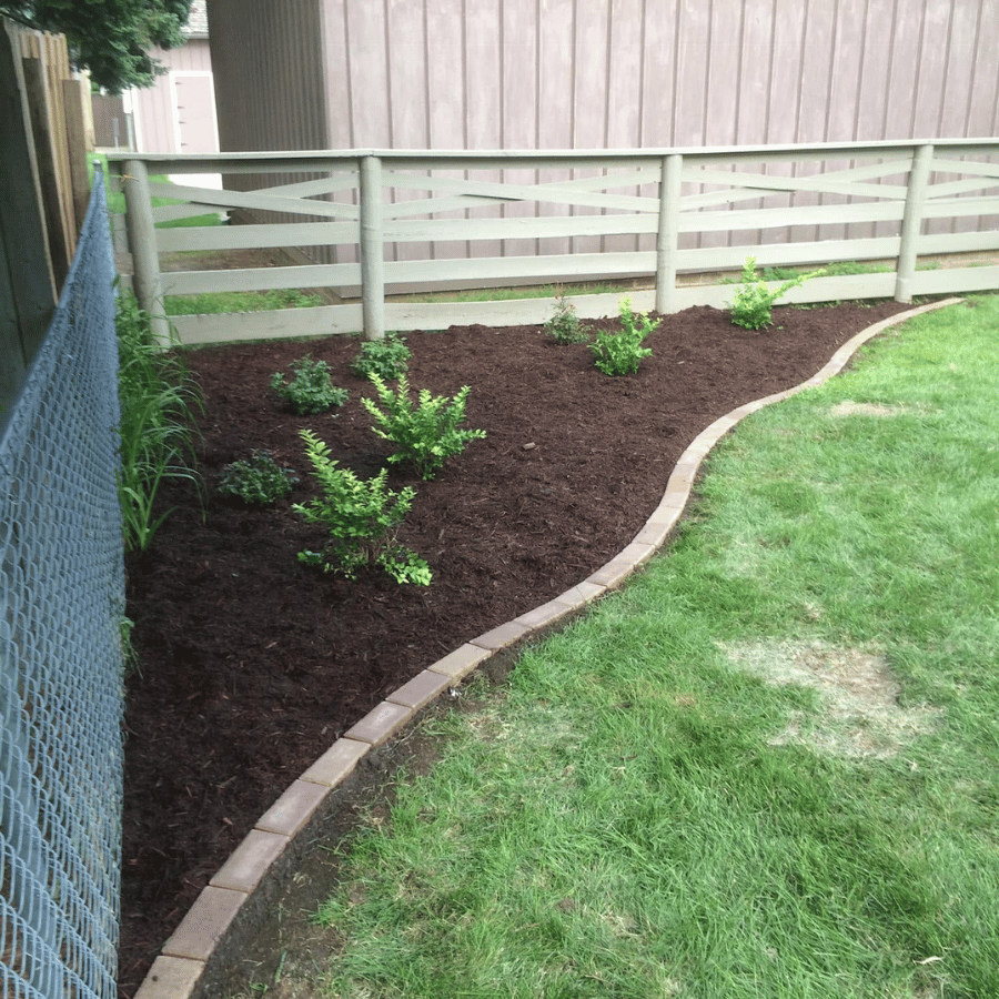Curved mulched garden bed with small shrubs bordered by pavers beside a grassy yard and white fence