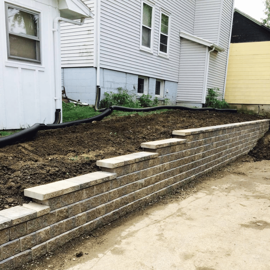Retaining wall with stone steps along a house, beside a dirt slope and driveway