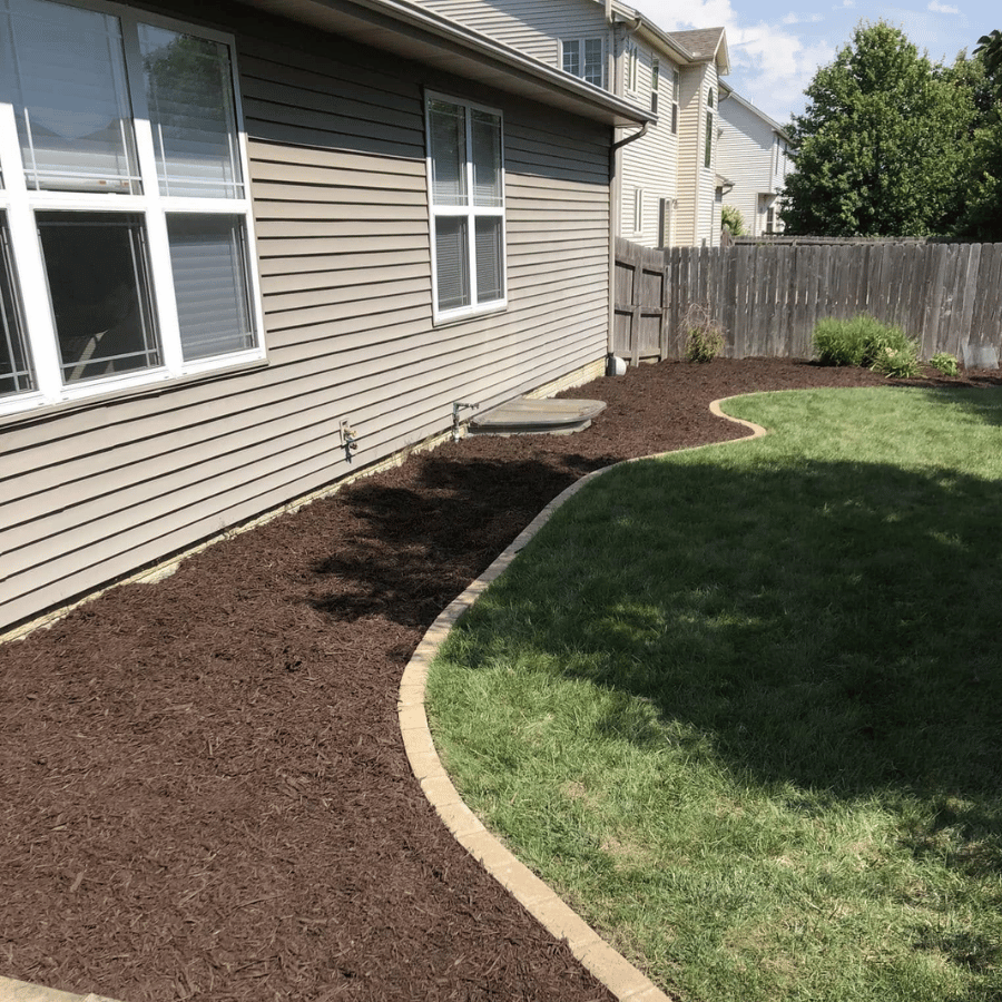 Mulched garden bed along a house’s side yard beside a curved green lawn