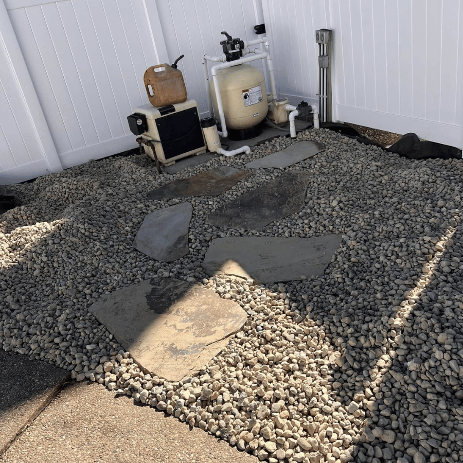 Backyard gravel area with stepping stones, a small pool filter system, and a tan tank against a white fence