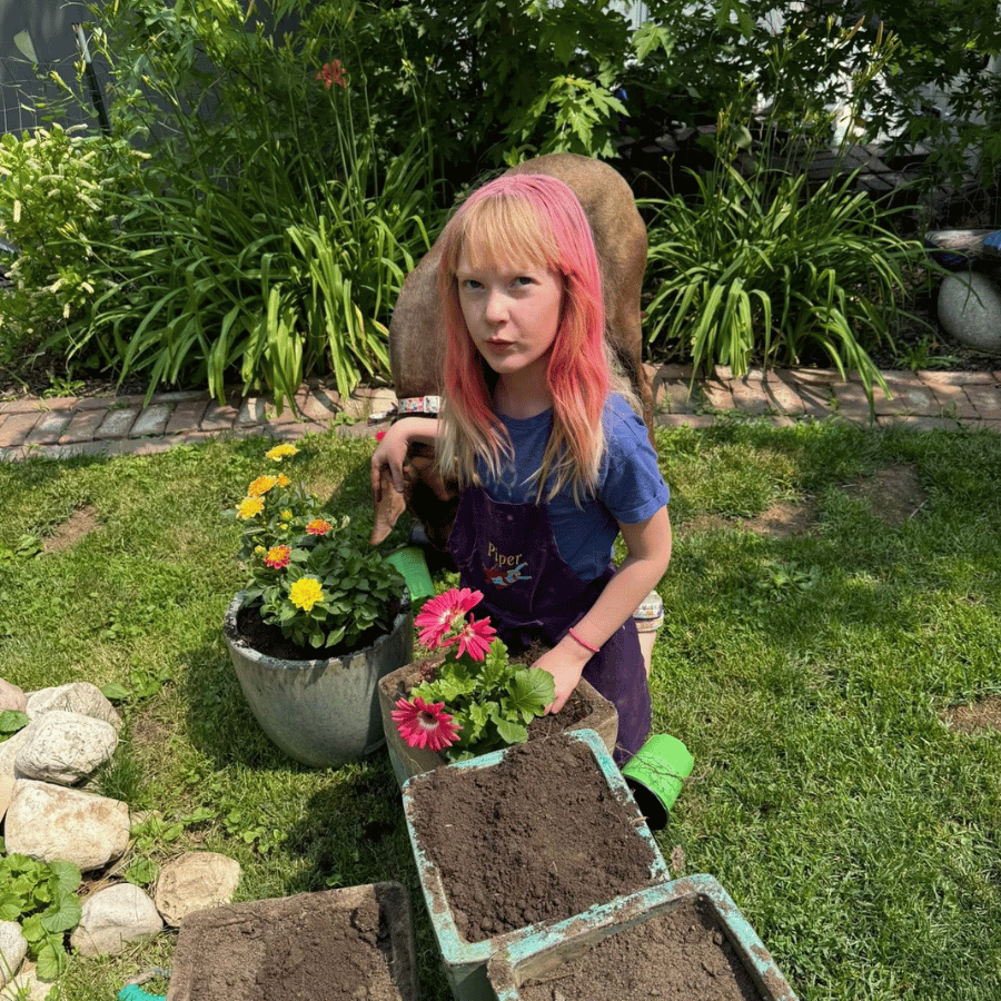 Child gardening in a backyard, holding pink flowers beside a small planter and pot of blooms.