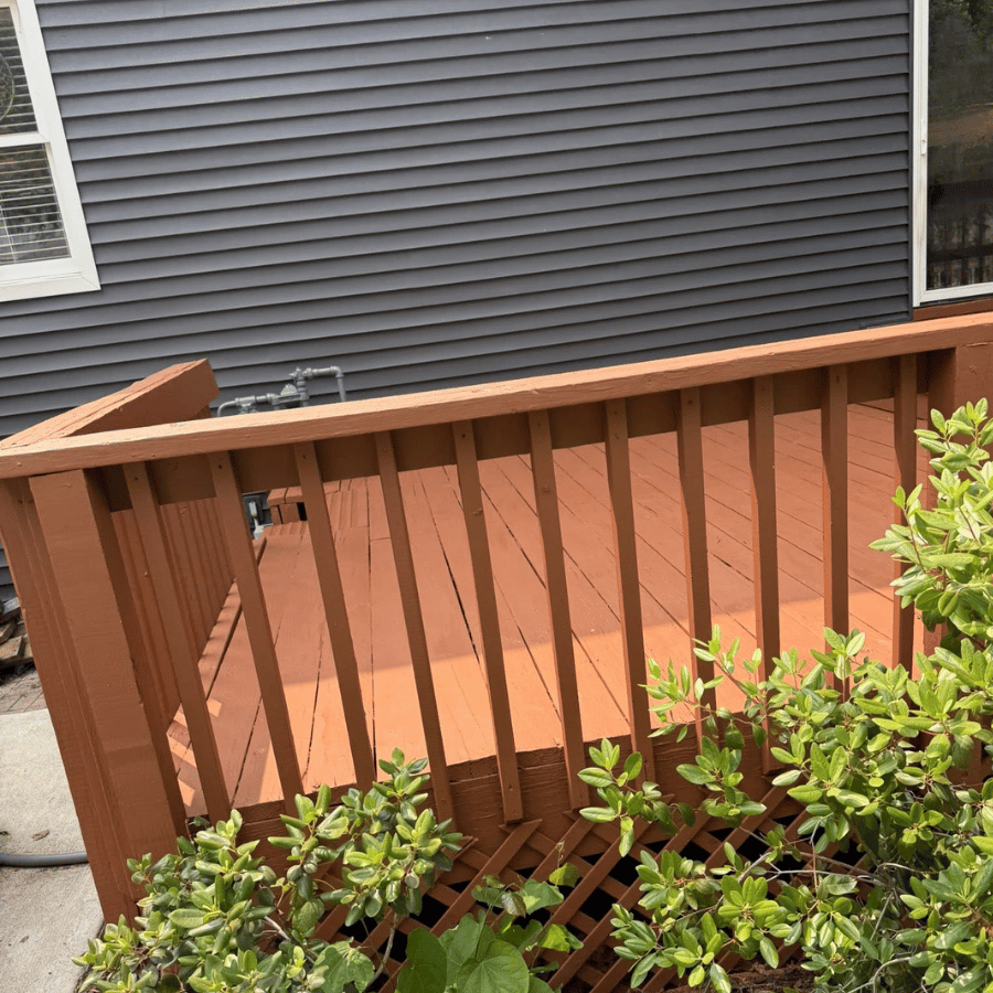 Wooden deck railing with brown posts beside gray house siding and green shrubs in the foreground
