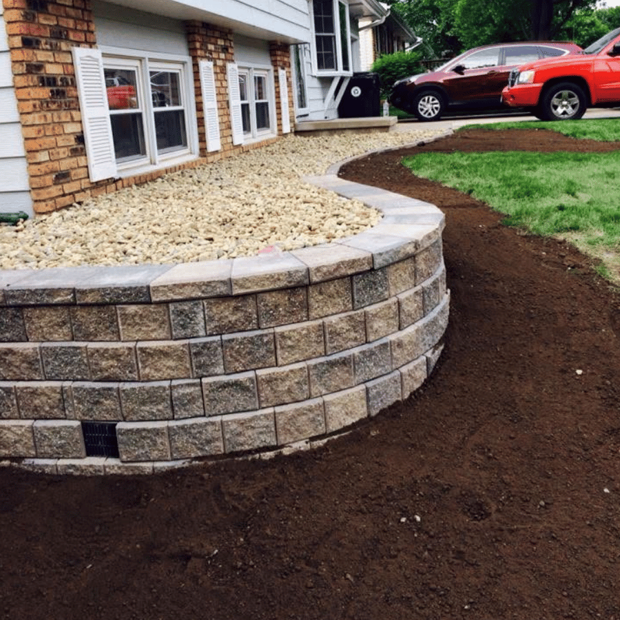 Curved stone retaining wall with gravel bed, bordered by fresh soil, adjacent to a house and parked red car.