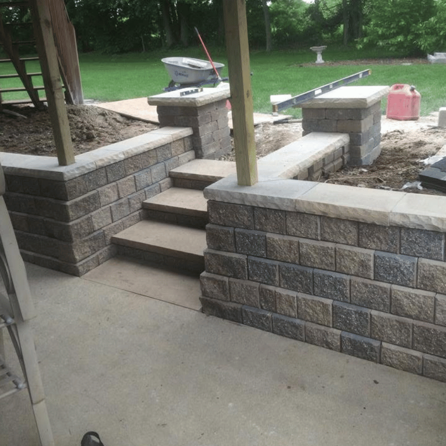 Stone steps lead to a garden with a wheelbarrow and tools, surrounded by a brick retaining wall under a wooden pergola.