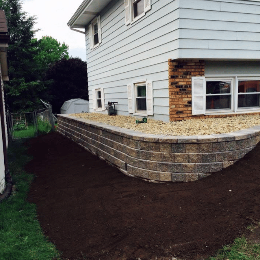 House with a new retaining wall and gravel landscaping, bordered by fresh soil, next to a green yard and a shed in the background.