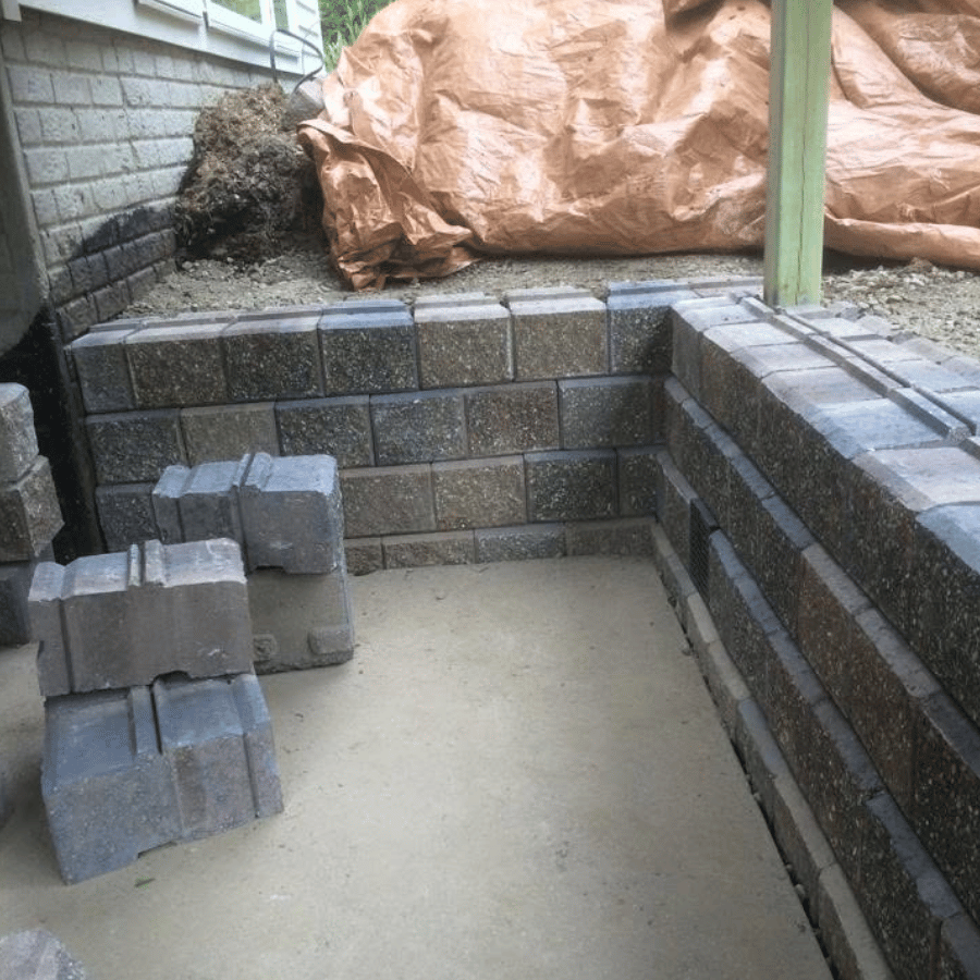 Stacked concrete blocks form a corner retaining wall on a patio. A tarp covers soil in the background.