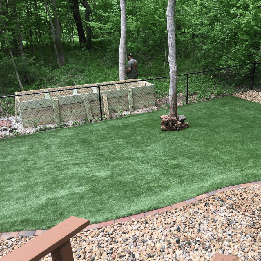 Man stands near wooden compost bin in lush backyard with artificial grass and trees, bordered by a black chain-link fence.